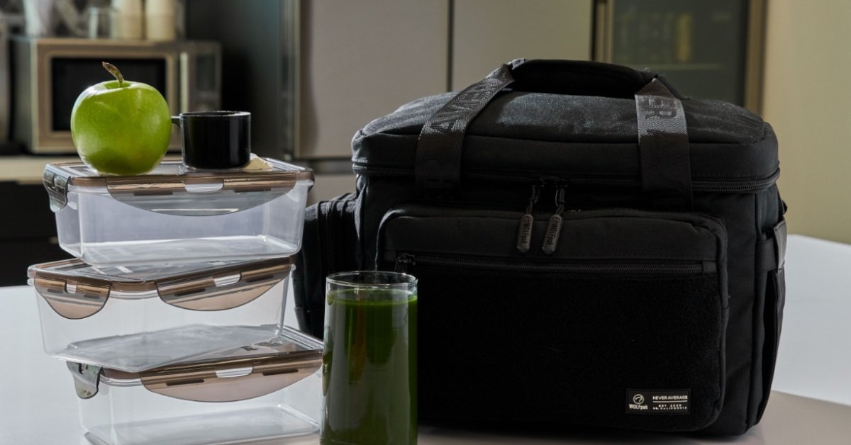 A black WOLFpak meal bag sits beside stacked food containers, a green apple, and a glass of green juice on a counter.