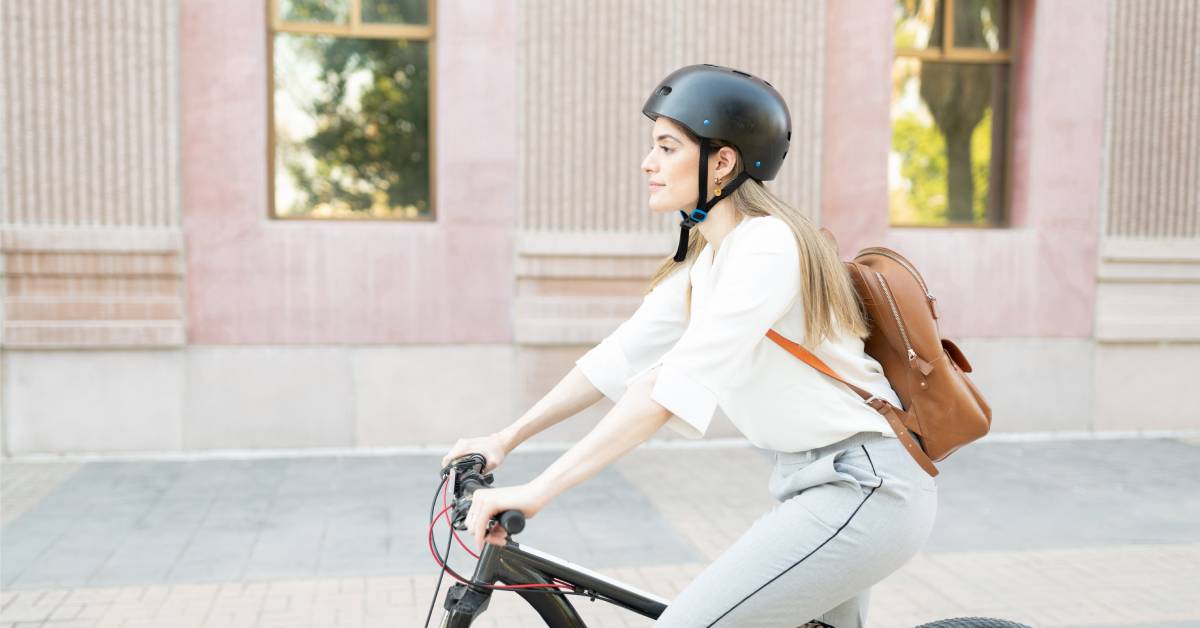 A woman in professional attire wearing a black helmet while riding a bike. She is also wearing a brown backpack.