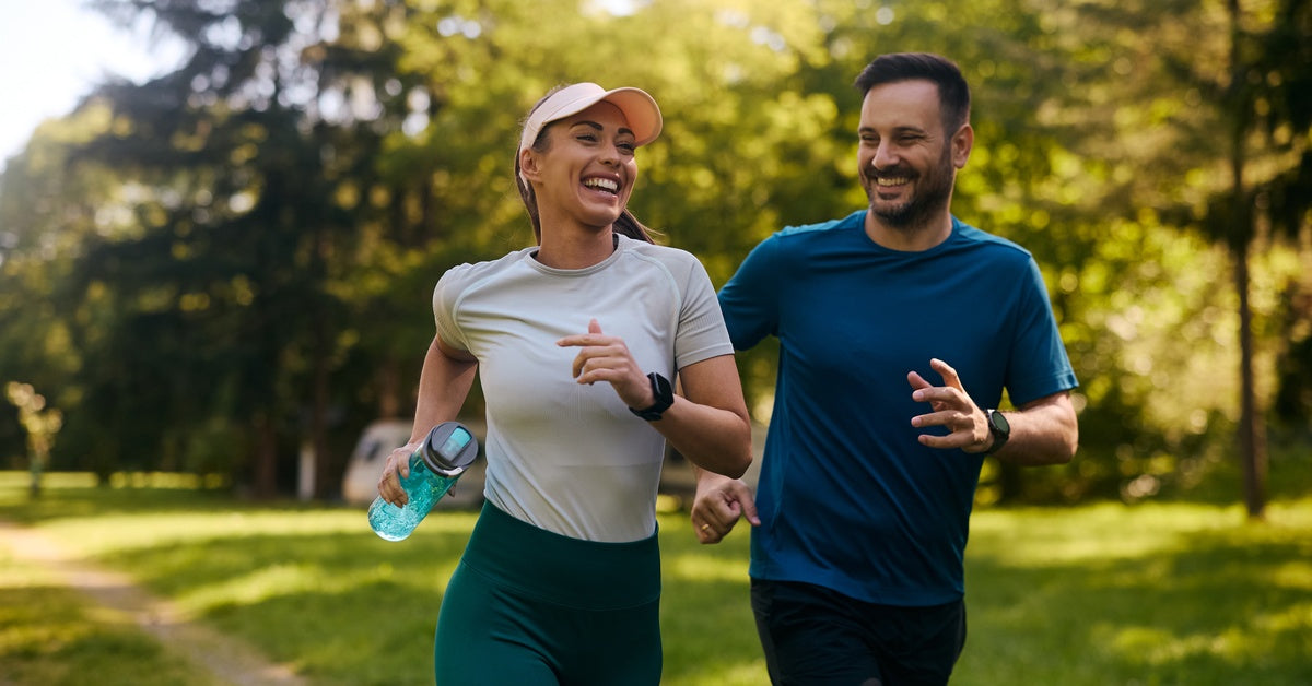 A man and woman are jogging next to each other outdoors in a wooded area while cheerfully smiling. The woman has a reusable water bottle.