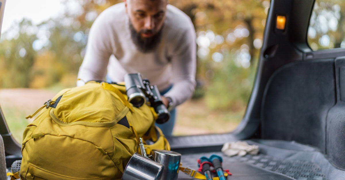 A man stands outside his car, gathering hiking essentials from the trunk and packing them into a yellow backpack.
