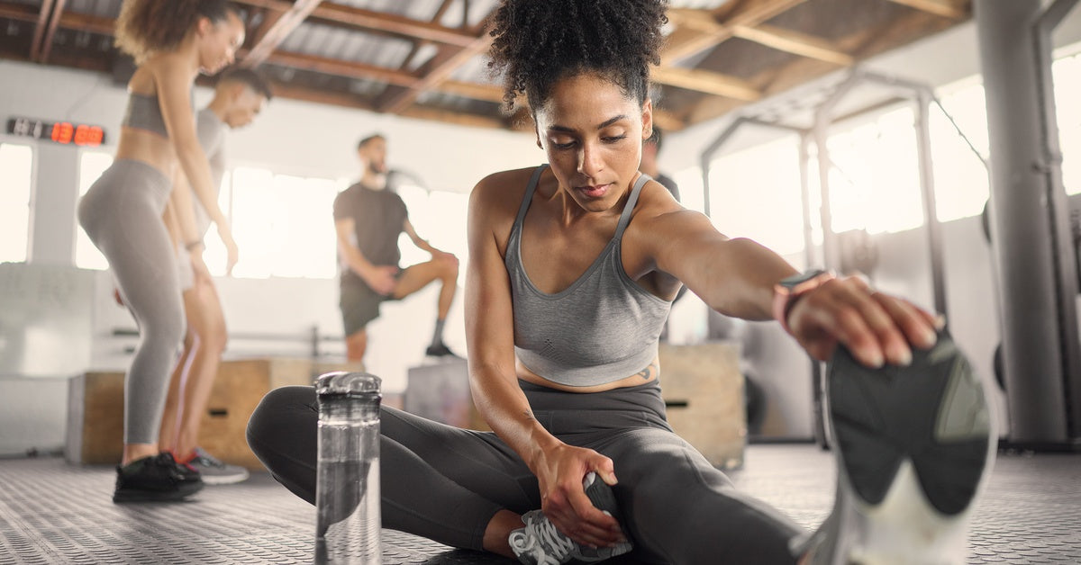 A woman sits on a padded gym floor and stretches with a water bottle beside her as others work out behind her.