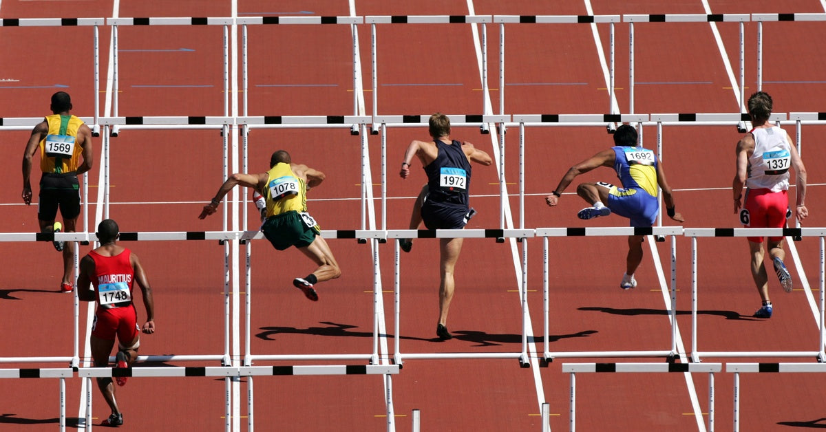 Six male athletes wearing bibs compete in a decathlon, actively jumping over hurdles on an outdoor track.