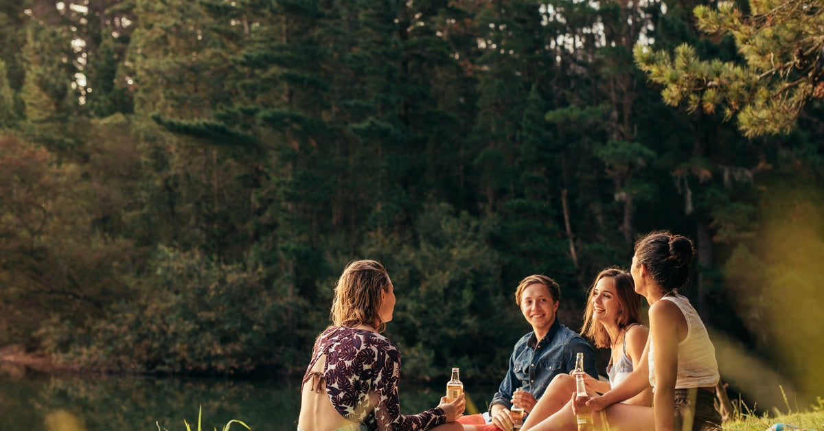 A group of friends smiling and laughing while having a picnic with food and drinks near a body of water on a sunny day.