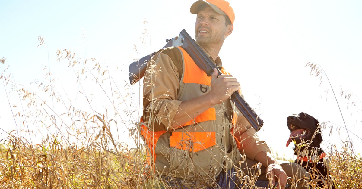 A hunter in high-visibility gear kneels in tall grass with a shotgun over his shoulder beside his alert hunting dog.