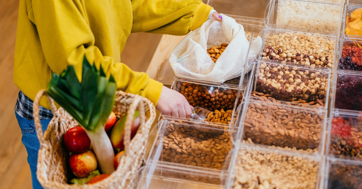 A woman in a yellow sweater uses a reusable bag to buy bulk nuts at a grocery store, with produce in a basket beside her.