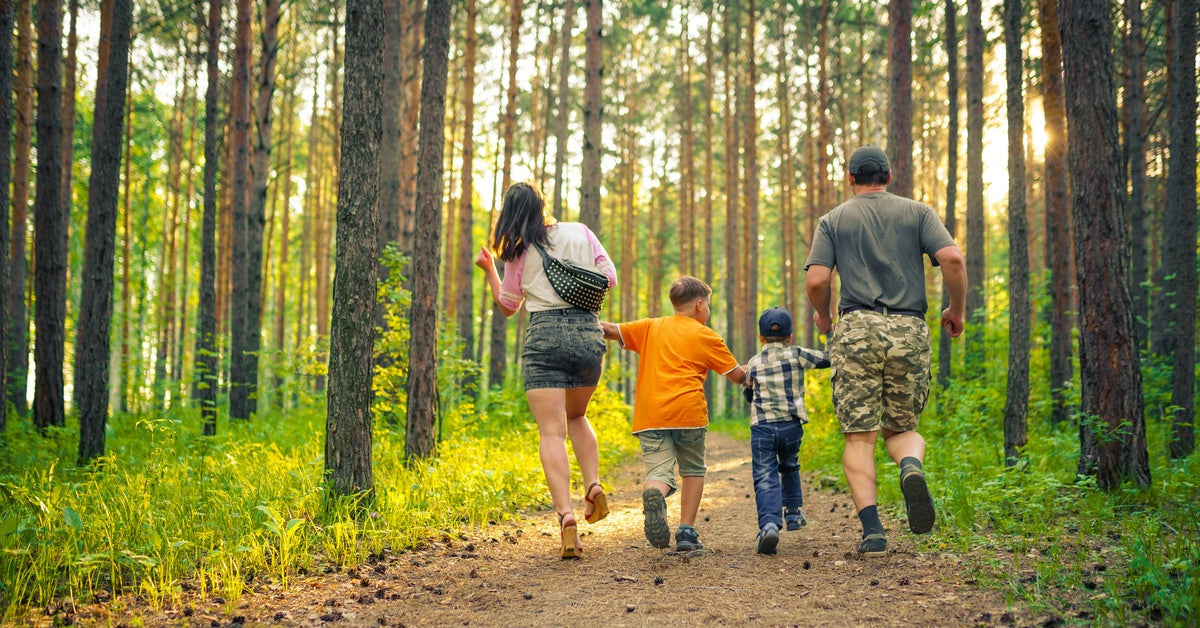 A women, man, and two young children dressed to hike hold hands while lightly jogging along a sunny forest trail.