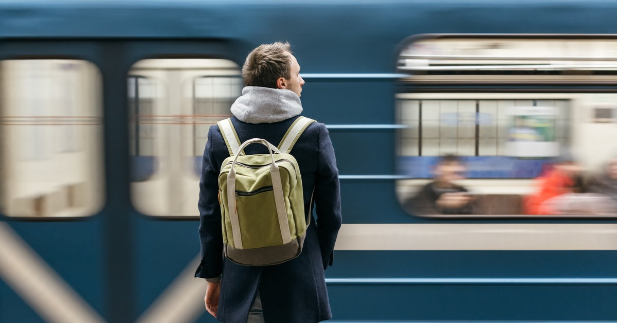 A man in a grey hoodie and blue coat with a backpack stands at a station facing a blue train speeding quickly by.