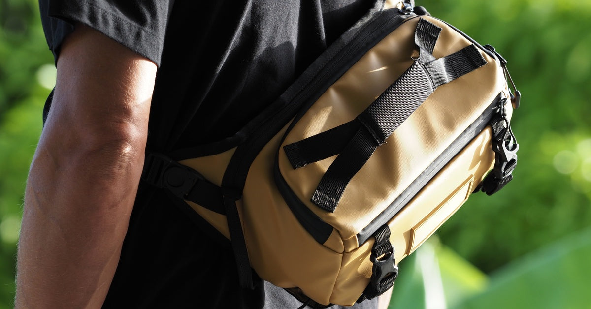 A person wears a tan crossbody bag with black straps and zippers, shown outdoors against a blurred green background.