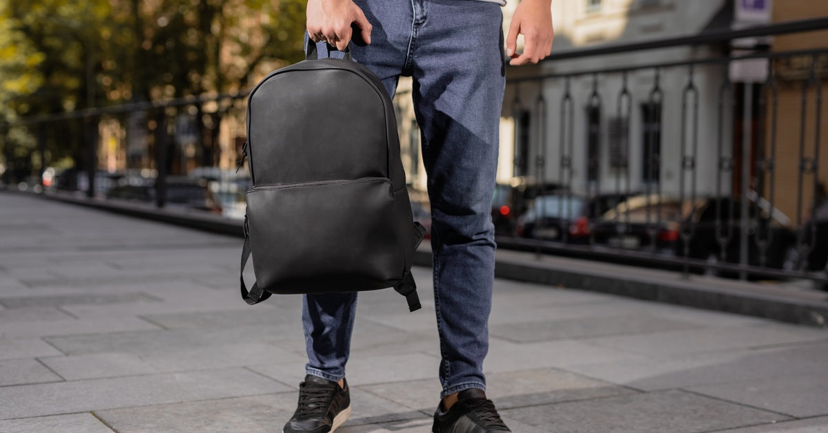A person walks along a city sidewalk holding a black backpack by the handle, with buildings and cars behind.