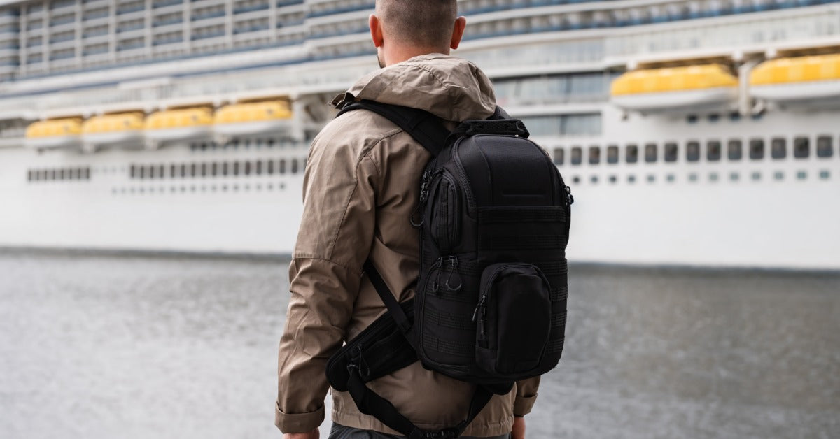 A man wearing a backpack is standing by the water, looking at a large cruise ship docked nearby in a harbor.