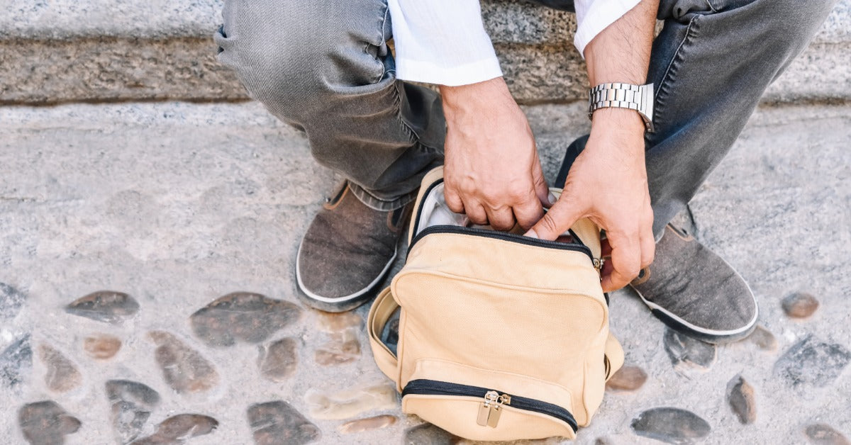 A person is opening a small beige backpack while seated on outdoor stone steps, viewed from above the bag.