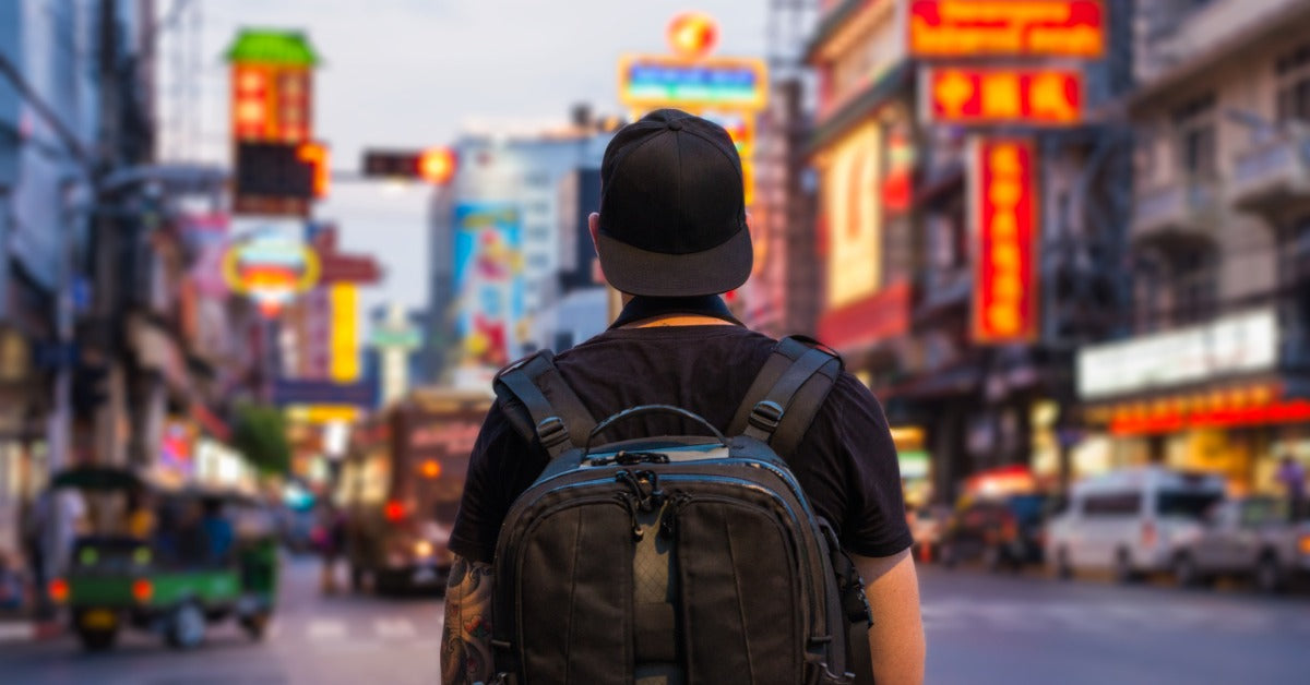 A traveler is wearing a backpack and stands in the middle of a busy city street, surrounded by colorful neon signs.