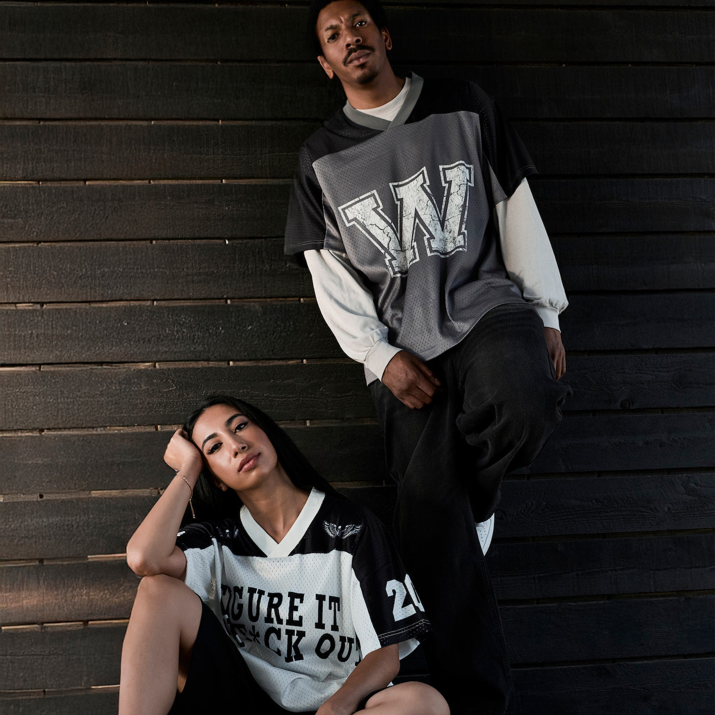 Two models posing in oversized graphic streetwear shirts against a dark wood plank wall. The man wears a gray, black, and white long-sleeve shirt with a large white 'W' graphic, and the woman wears a black and white V-neck graphic shirt.