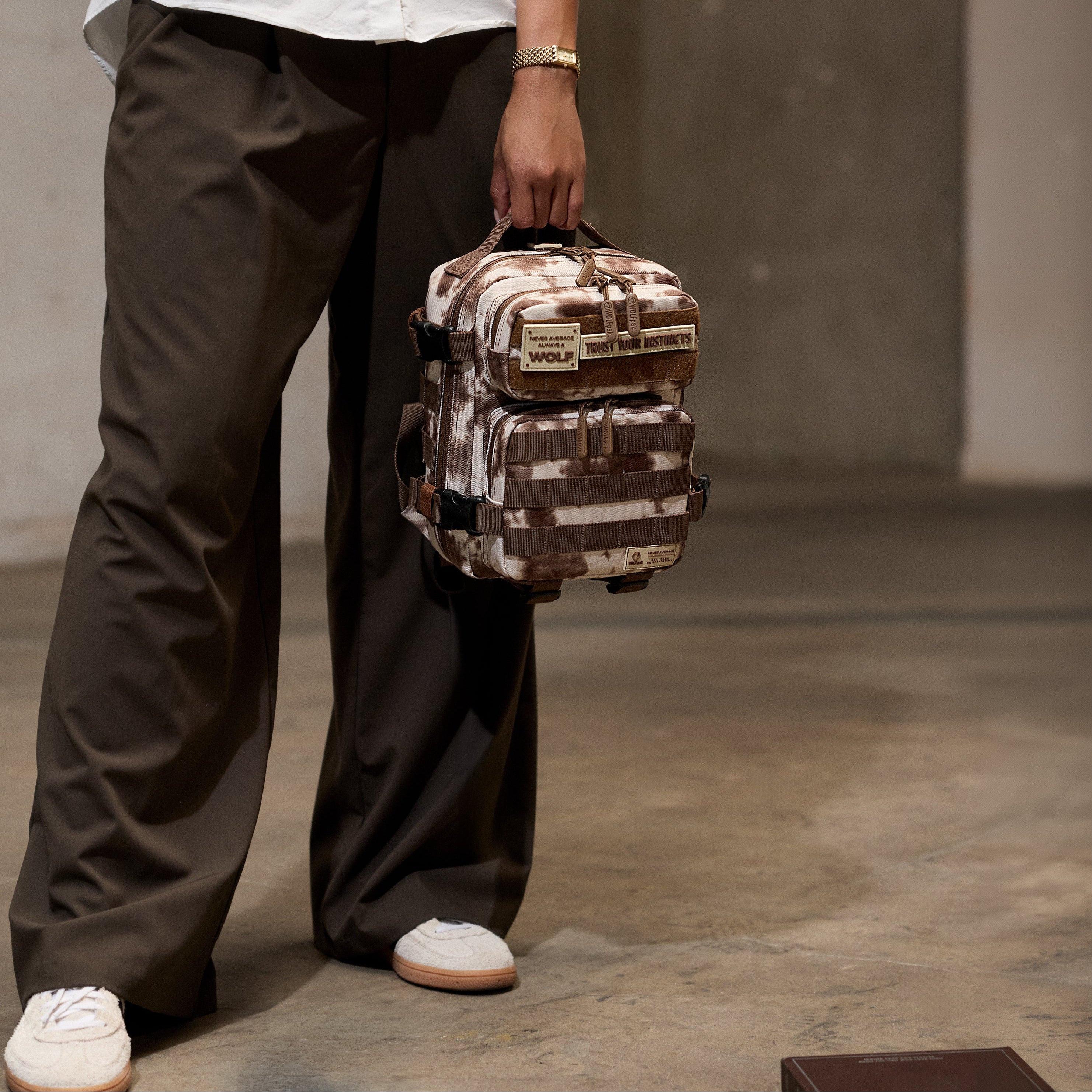 A person wearing a white button-up shirt, dark brown wide-leg pants, and white sneakers holds a small tactical-style backpack with a brown and beige camouflage pattern in a minimalist concrete room.