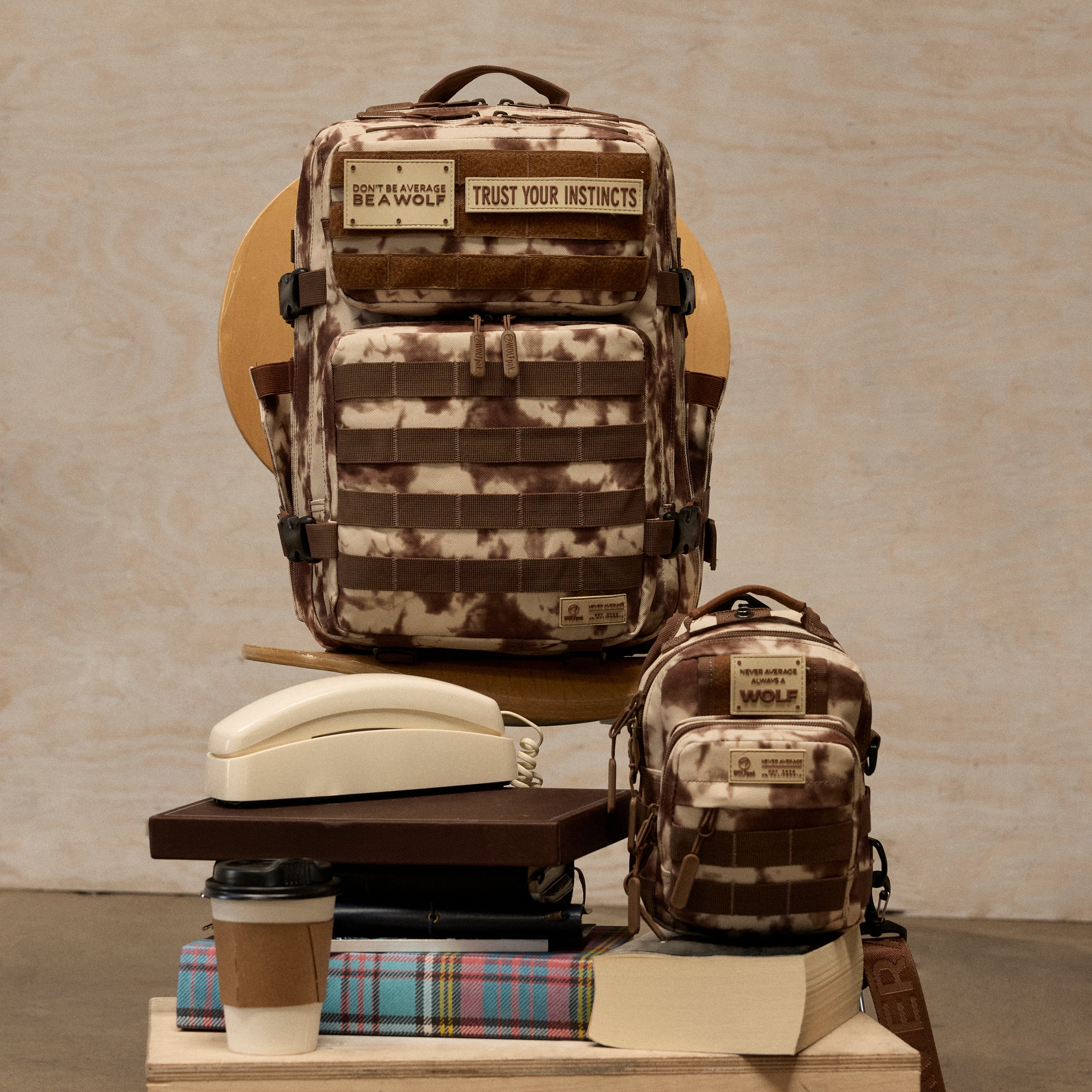 Two brown and beige camouflage backpacks from The Bear & Wolf Collection are displayed on a wooden stand with books, a retro phone, and coffee cups. The larger backpack features a front utility pocket with a patch reading 'TRUST YOUR INSTINCTS' and a top patch reading 'BEAR WOLF'.