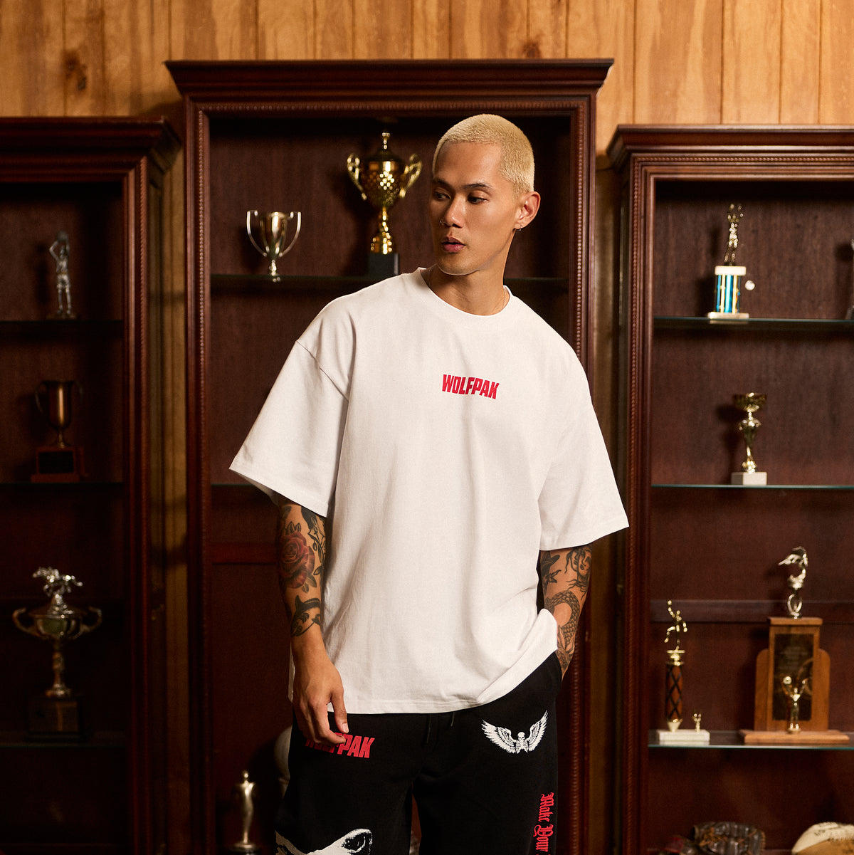 A man with a buzz cut stands in front of wooden trophy shelves wearing an oversized white t-shirt with 'WOLFPAK' printed in red on the chest, paired with black shorts.