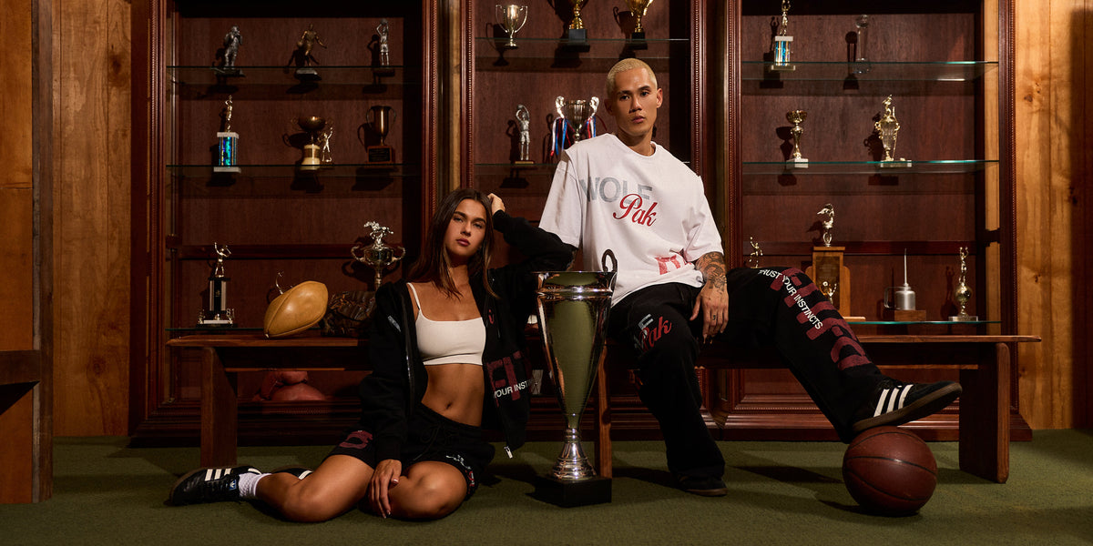 A man and a woman pose in a trophy-filled room. The man wears a white graphic T-shirt and black pants, sitting next to a large silver trophy with a basketball on his leg.