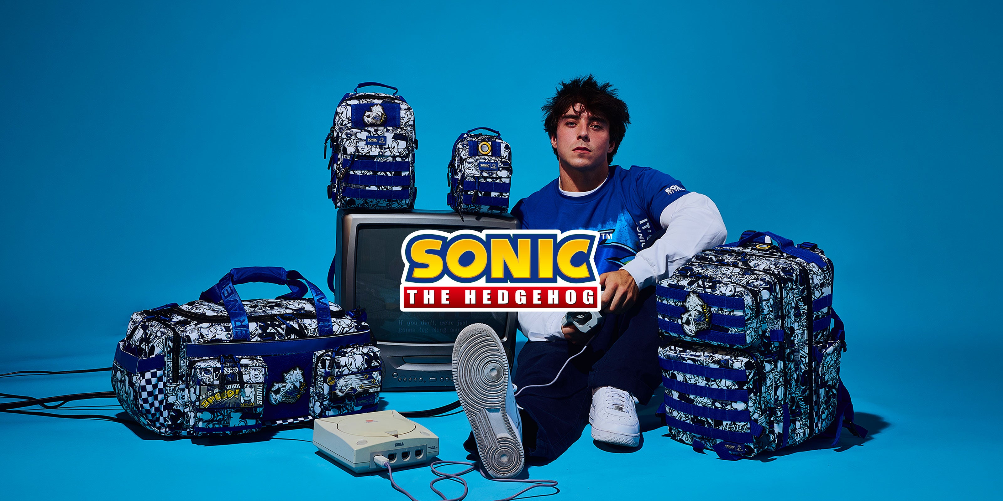 A person sits on a bright blue studio floor surrounded by multiple Sonic the Hedgehog-themed backpacks, duffel bags, a retro TV, and a console. The person is wearing a blue and white Sonic jersey, and the products feature Sonic's signature blue and white camouflage pattern.