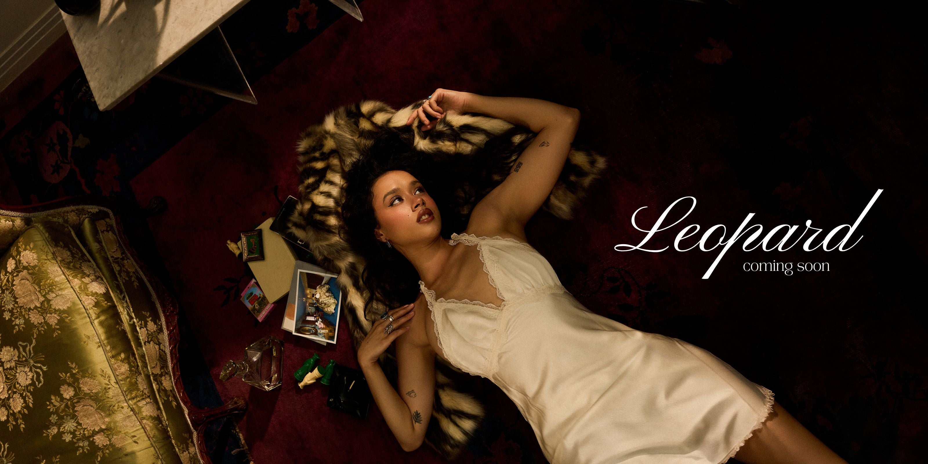 A woman reclines on a dark red patterned rug wearing a white satin slip dress, with a leopard print throw blanket beside her. The word 'Leopard' appears in white script on the right side, with 'coming soon' below it.