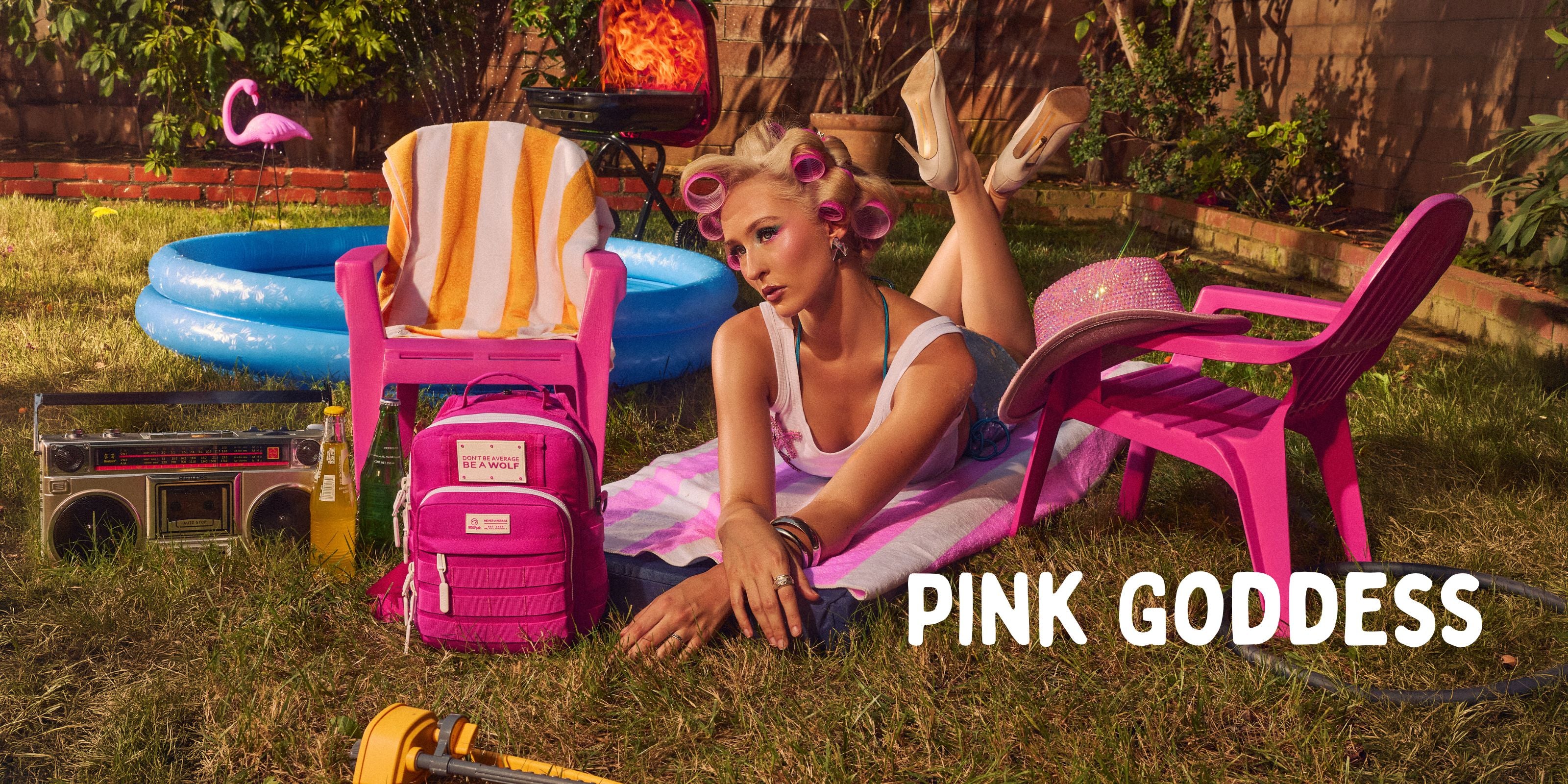 A woman reclines on a pink lawn chair in a backyard setting, wearing a pink and white striped bikini and a pink backpack. A pink striped towel sits on a pink chair, with a blue pool, boombox, and flamingo decoration nearby.