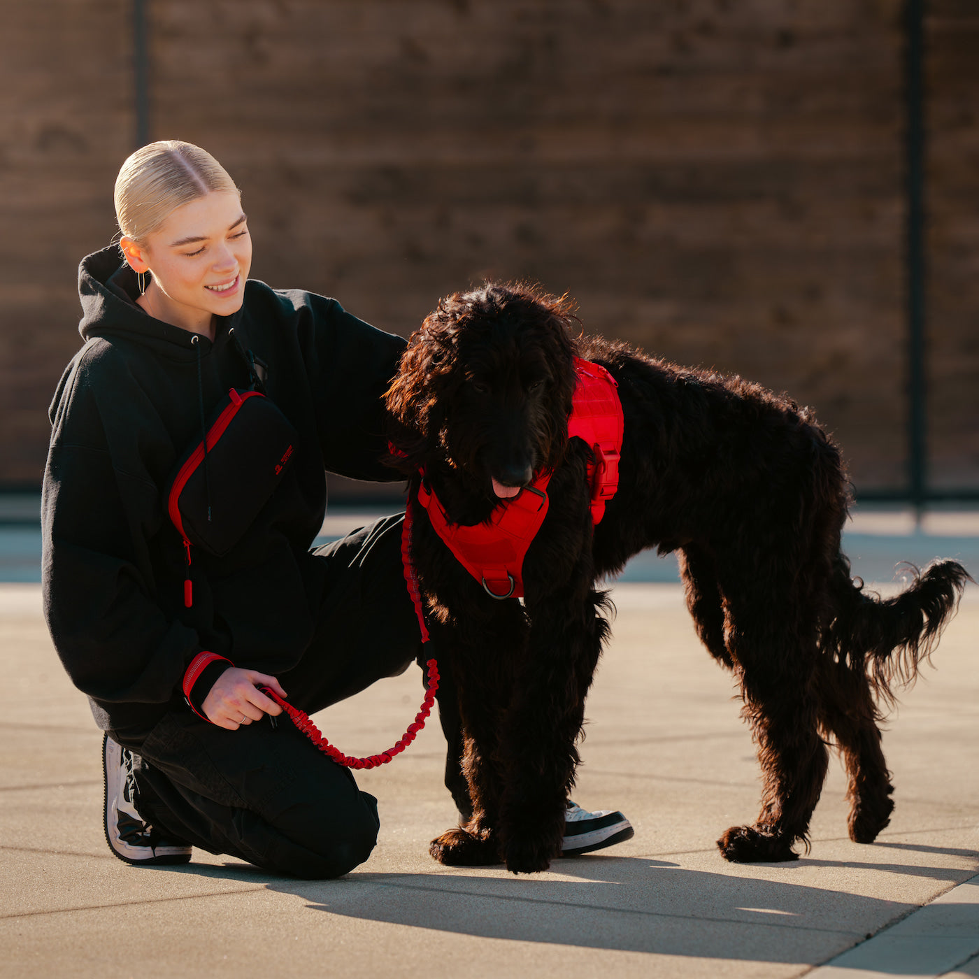 A woman kneels beside a black curly-haired dog outdoors, wearing a black hoodie with red trim and holding a red leash. The dog wears a red harness.
