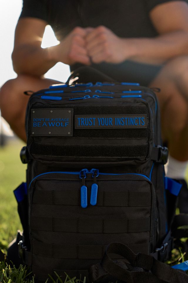A black tactical backpack with blue accents sits on grass while a person adjusts the straps behind it outdoors.
