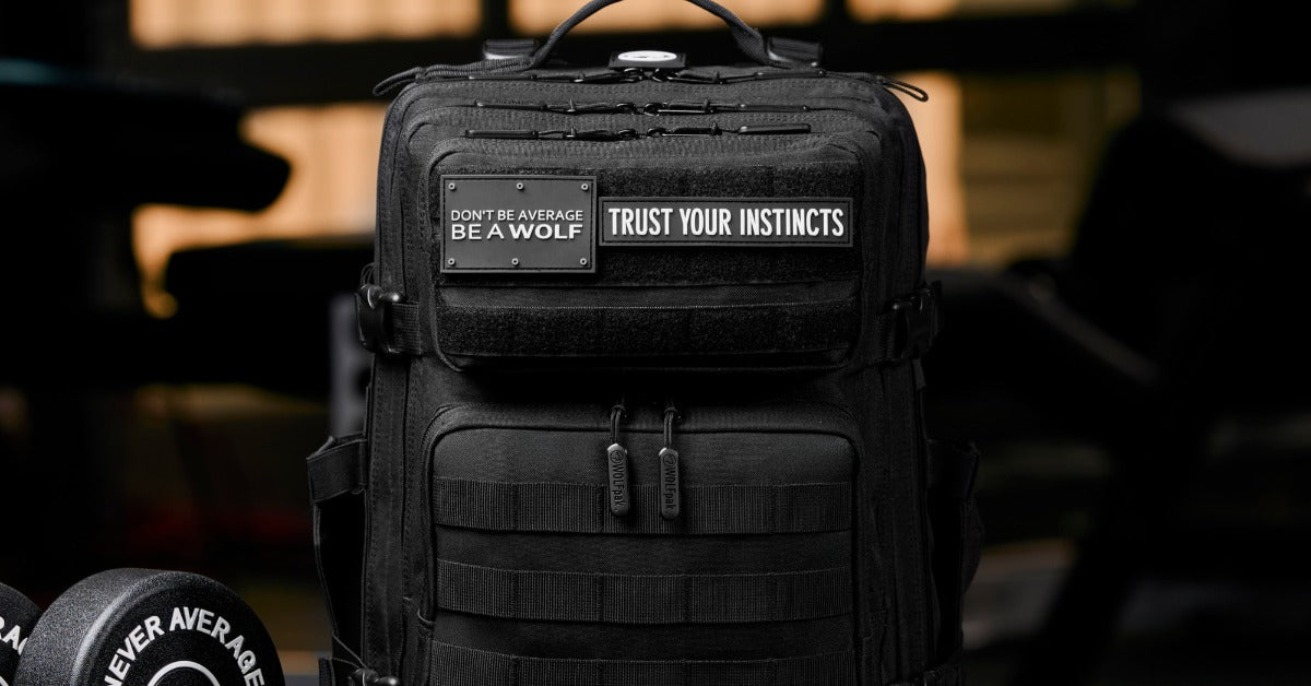 A black 25L tactical backpack with MOLLE webbing and motivational patches sits upright beside gym equipment.