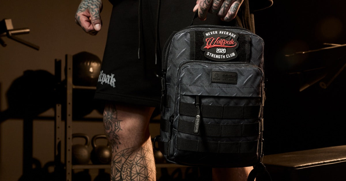 A tattooed person holds a compact black WOLFpak backpack with MOLLE webbing and a Never Average patch in a gym.