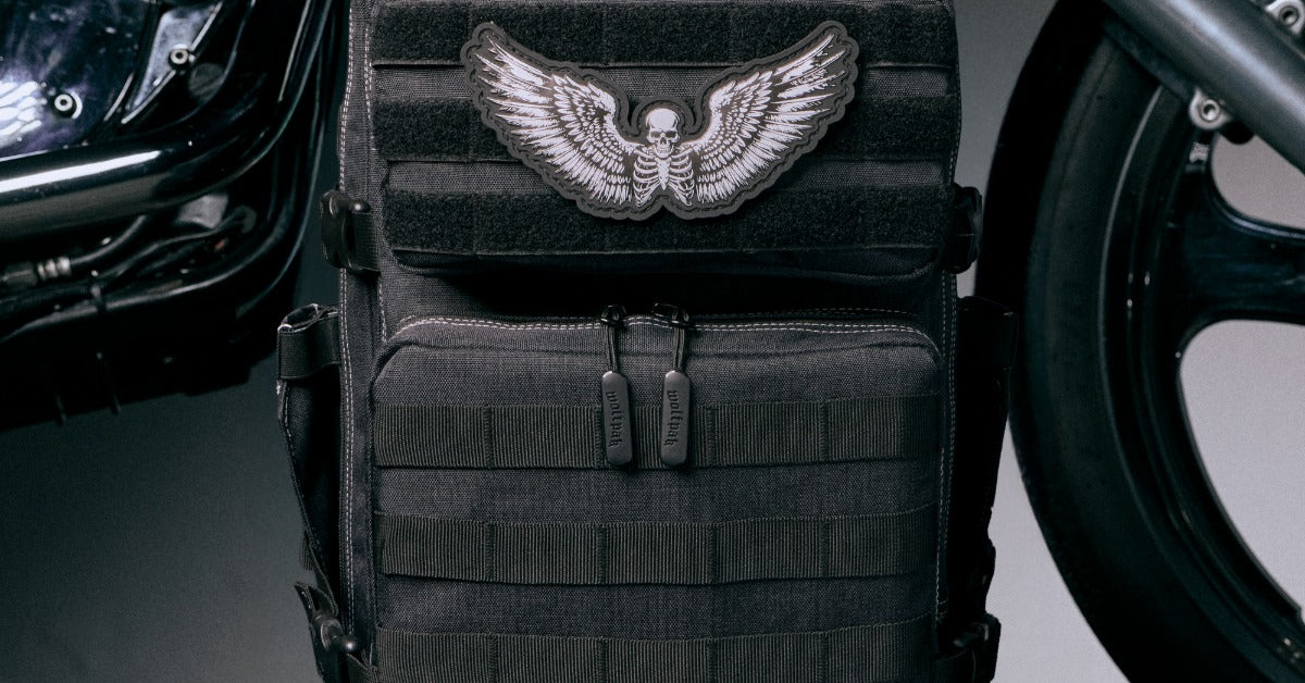 A black tactical backpack with MOLLE webbing and a winged skeleton patch stands between two black motorcycles.