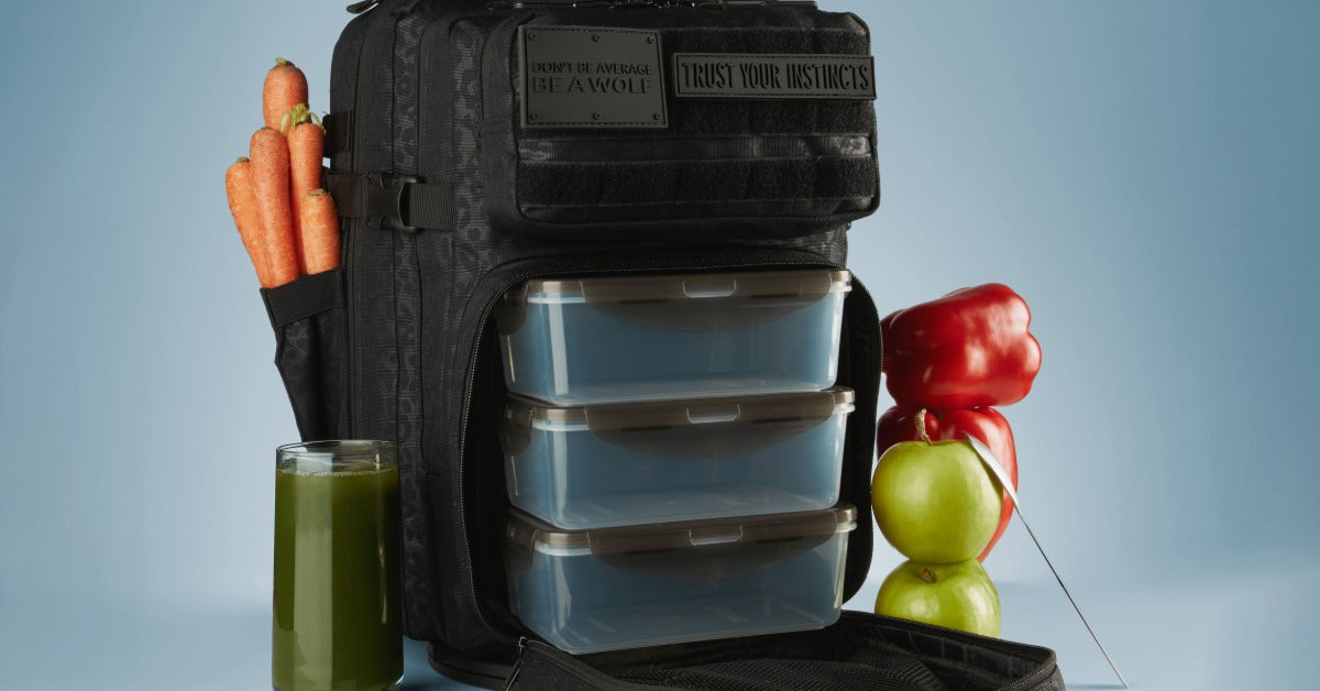 A black WOLFpak meal prep backpack holds three stacked food containers beside carrots, peppers, apples, and green juice.