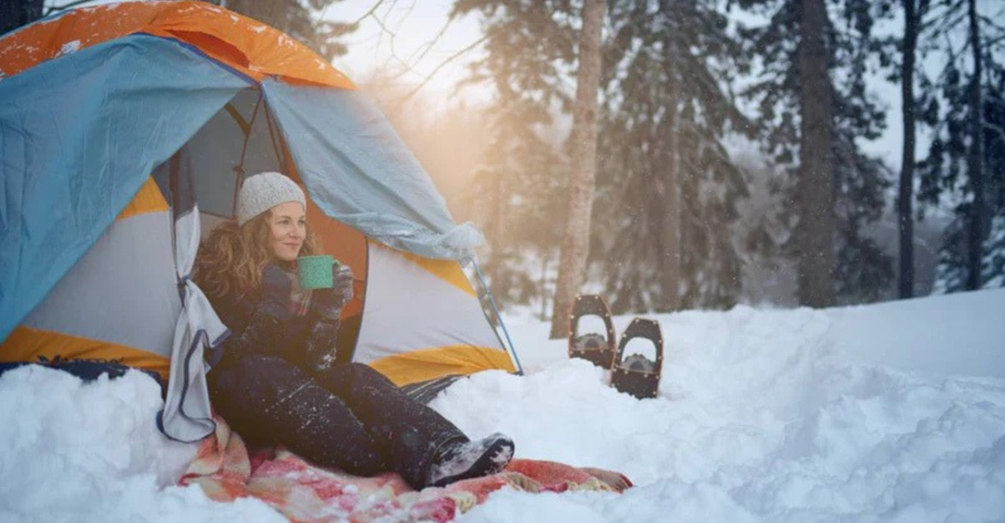 A woman dressed in winter gear sitting in a tent surrounded by snow and trees. She is holding a green mug.