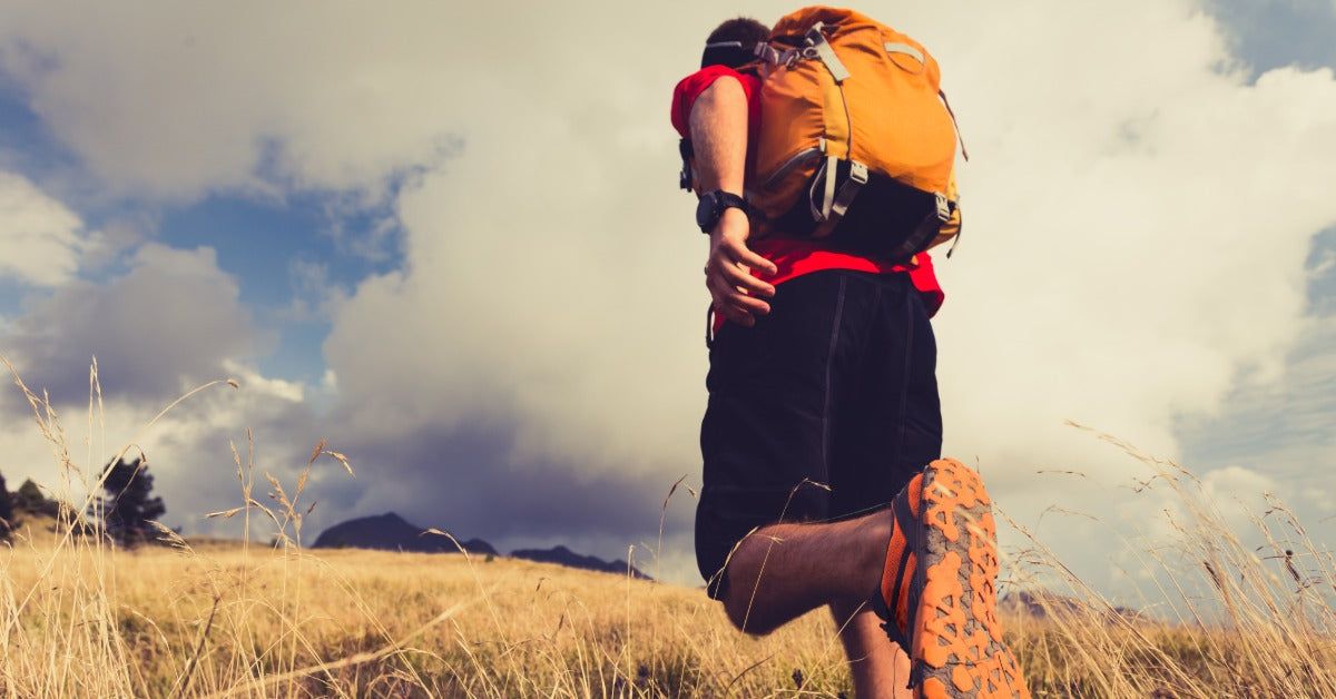 A hiker on a grassy trail hiking up a hill. He is wearing athletic shoes and a hiking backpack on his back.