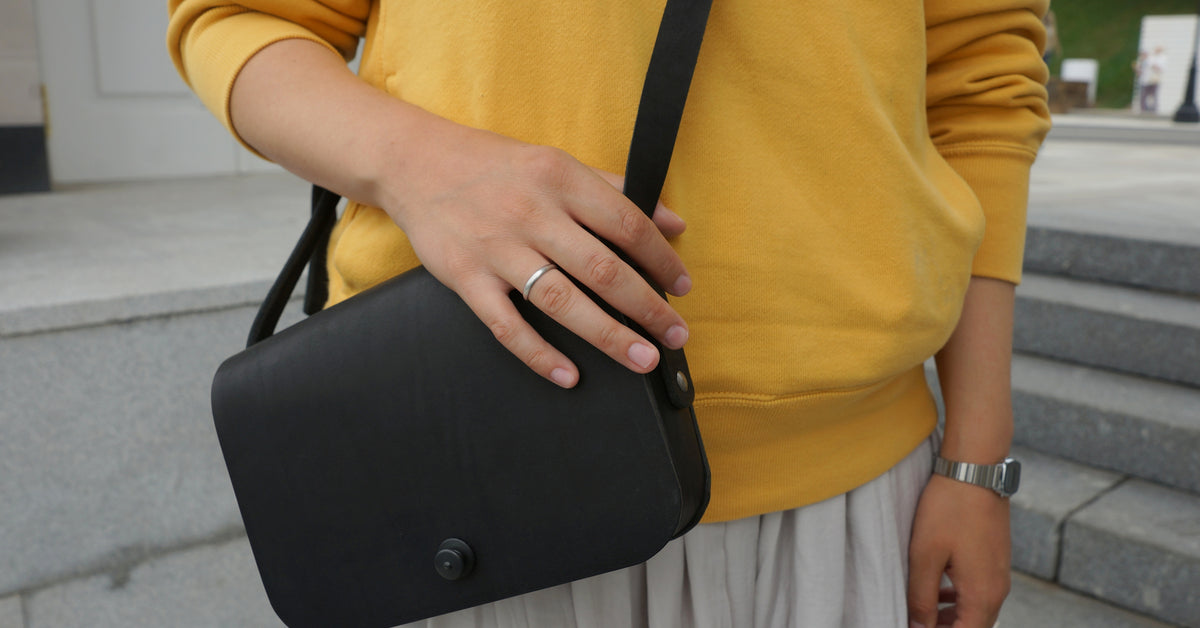 A woman is standing outside wearing a yellow sweatshirt and a white skirt. She has a black crossbody bag on her shoulder.