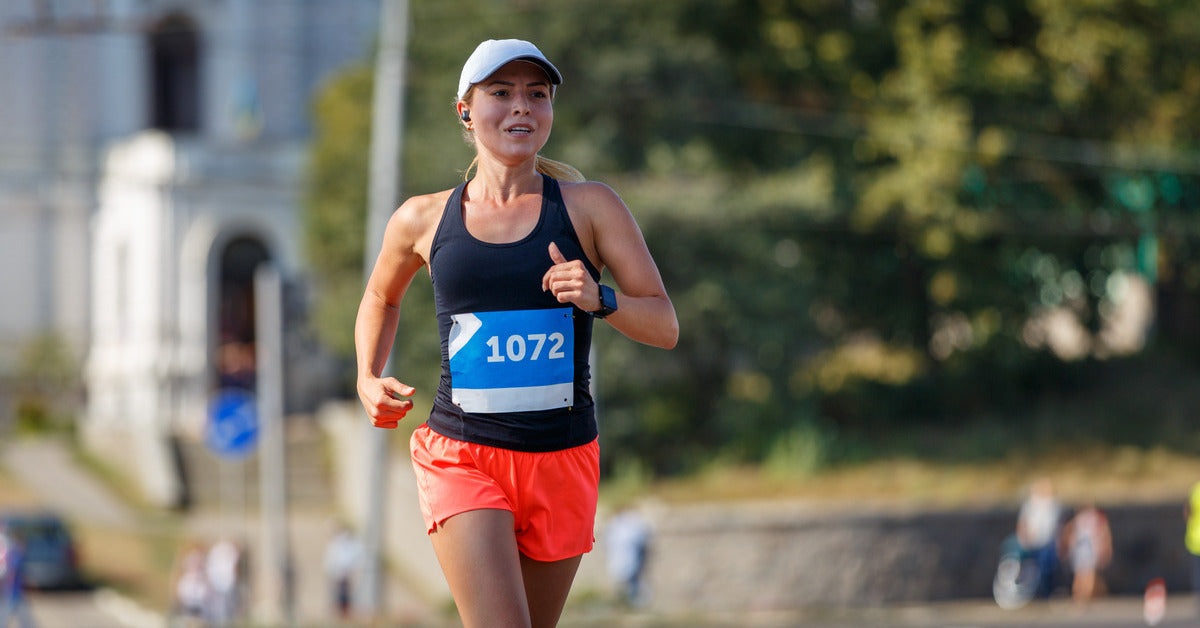 A woman is running outside. She is wearing shorts, a tank top, a hat, and high socks. She has a race bib pinned to her shirt.