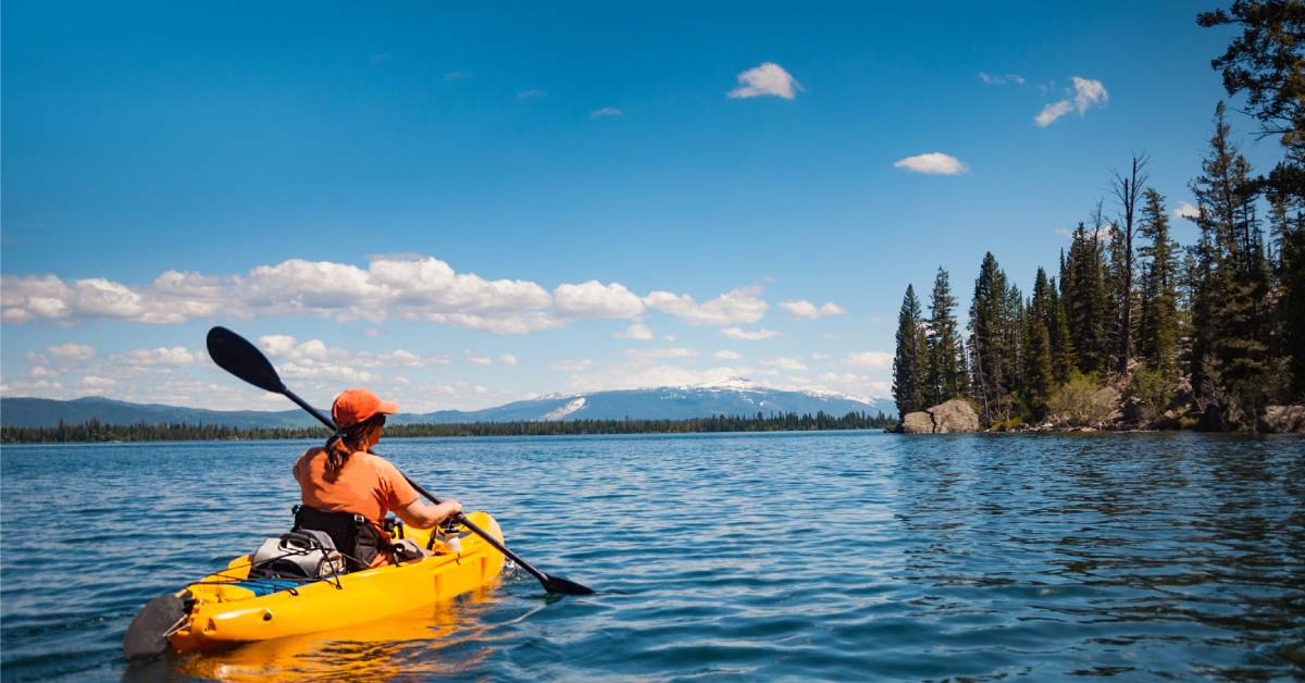 A woman kayaking on a lake surrounded by trees and mountains on a sunny day. She has a backpack strapped to the kayak.