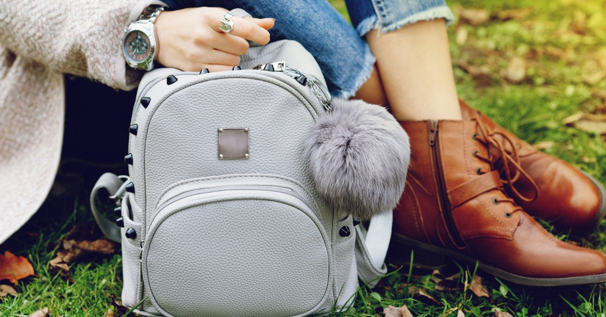 A stylish woman is sitting outside on the grass. She is holding a gray backpack with stud details and a fluffy keychain.