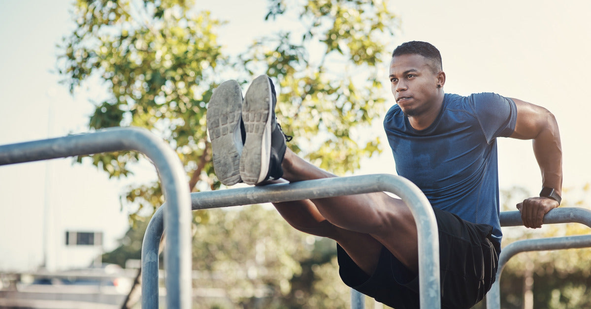 A man is wearing workout attire and balancing himself between two metal bars outside. He has a focused look on his face.