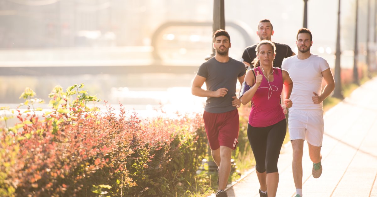 Four people jogging down a city sidewalk in daylight, with flowers beside them and city lights lining the path.