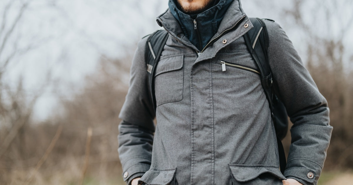 A younger man wearing a layered gray winter coat with a hood, carrying a black backpack while outdoors in cold weather.