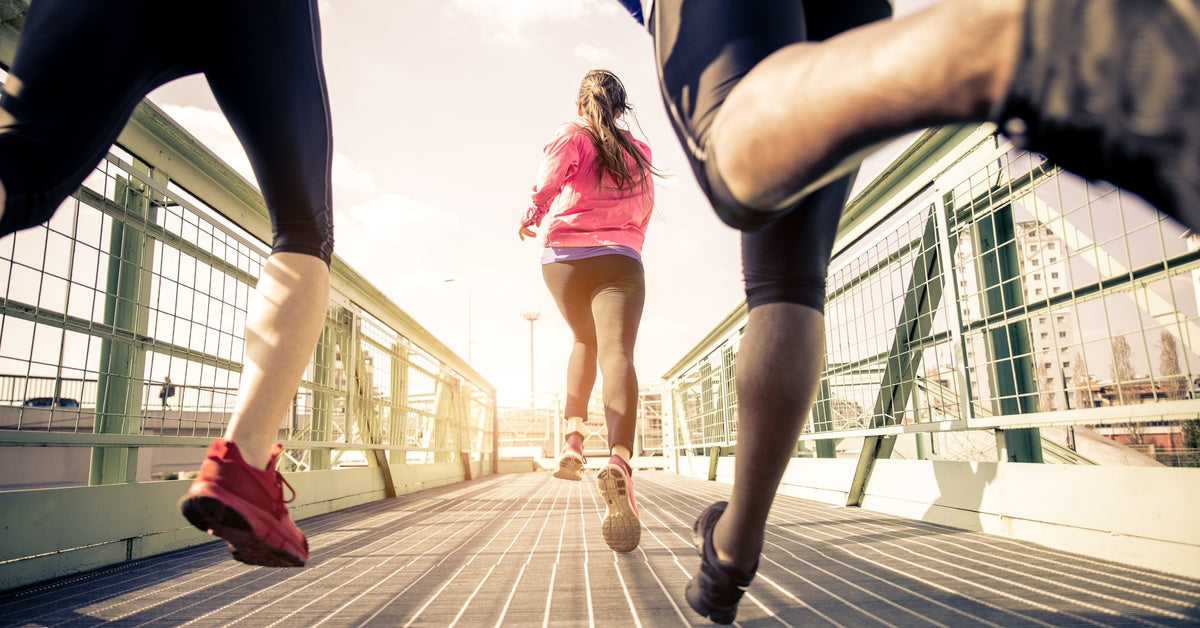 Three joggers wearing workout clothes run across a green metal bridge in a city as the sun rises in front of them.