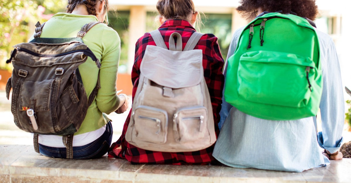 Three teenagers—two girls and one boy—sit on a stone ledge at school, each wearing a different type of backpack.