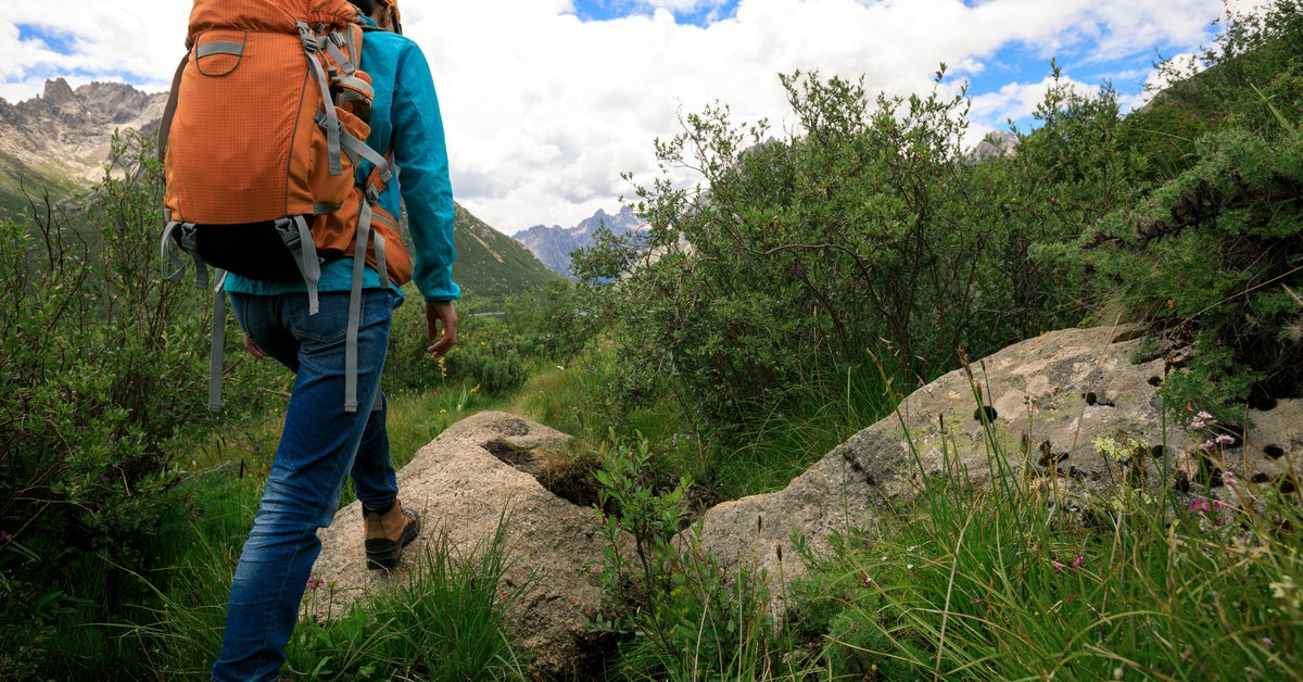 A thin young person wearing jeans, a jacket, and an orange hiking backpack hikes across rugged terrain with mountains nearby.