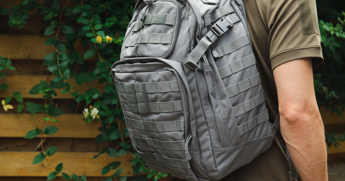 A man in military-style clothing stands outdoors near a fence and greenery with a gray tactical backpack on his back.