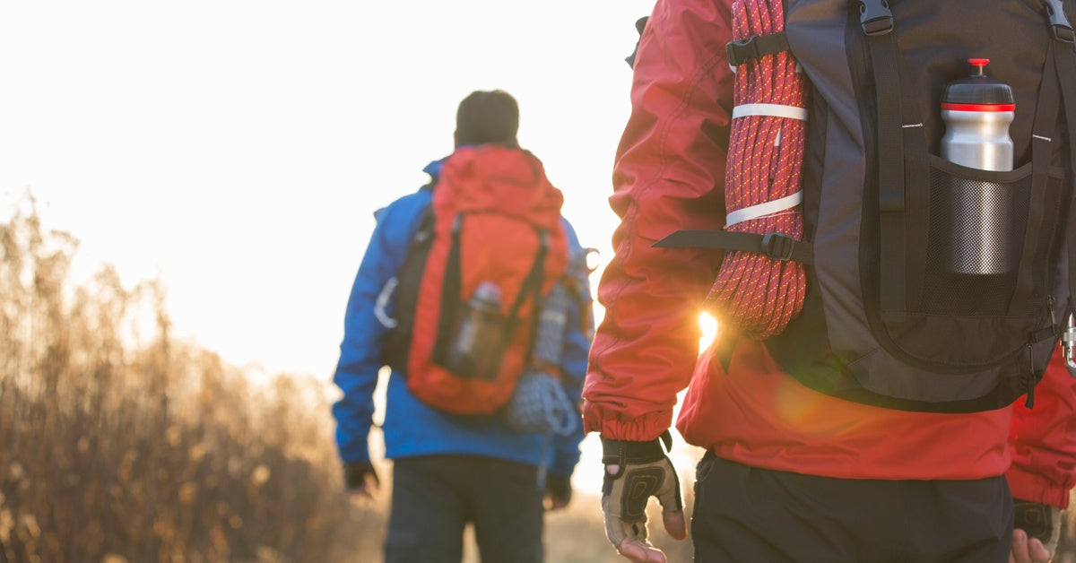 Two winter hikers in heavy coats, beanies, and gloves walk with backpacks that hold a rope and a water bottle.