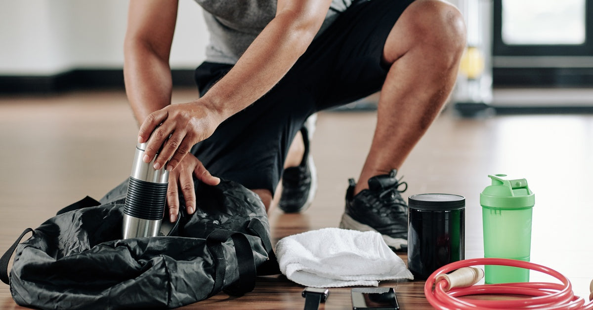 A man in gym clothes kneels and packs a black duffel bag with a water bottle beside workout gear on the floor.