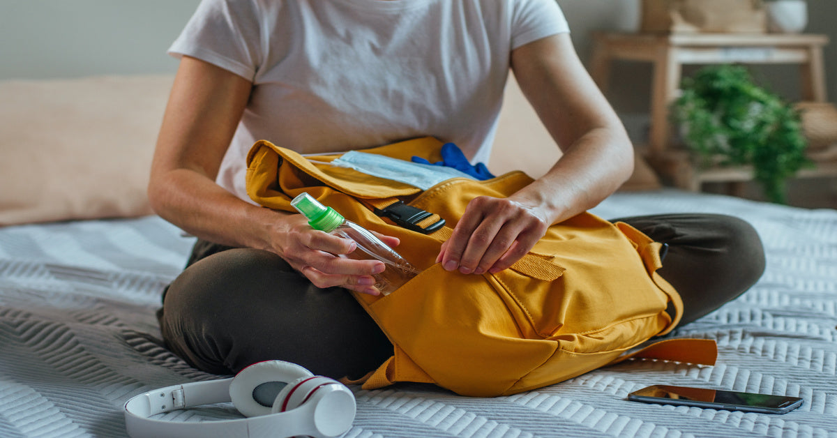 A woman sits on her bed packing a yellow backpack with daily essentials, including headphones and sanitizer.