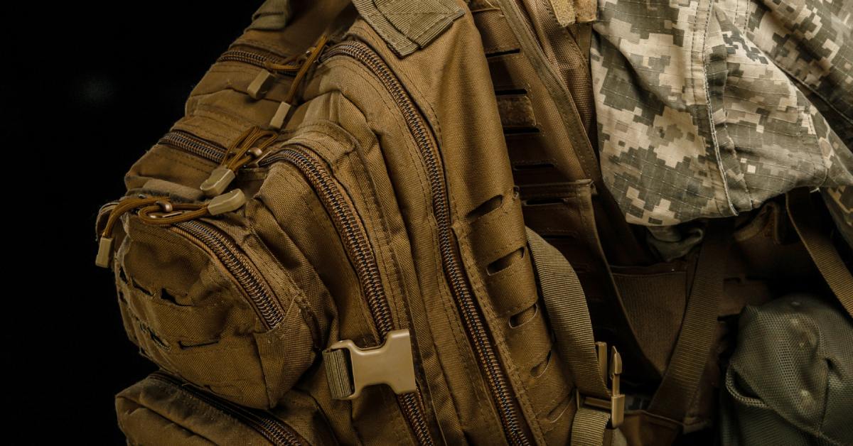  Close-up of a tactical military backpack with multiple zippered compartments worn over a camouflage uniform.