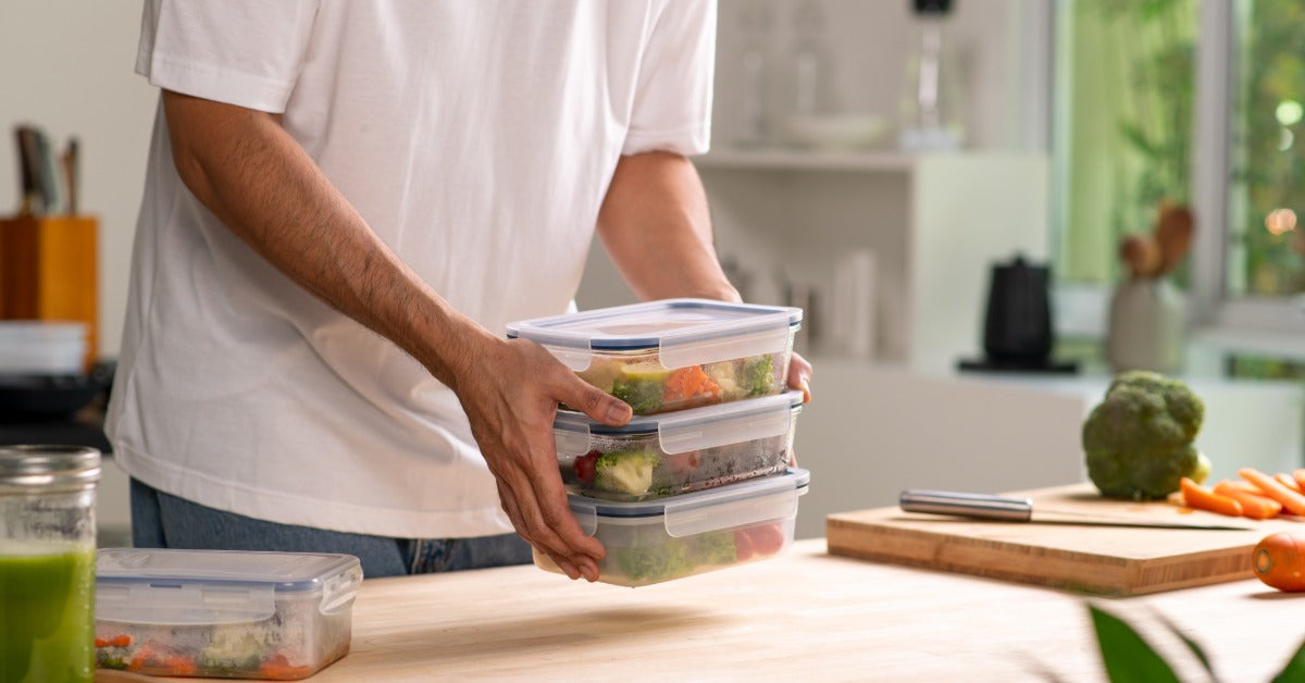 A man wearing a white t-shirt in a kitchen stacks clear plastic containers filled with prepared healthy meal portions.