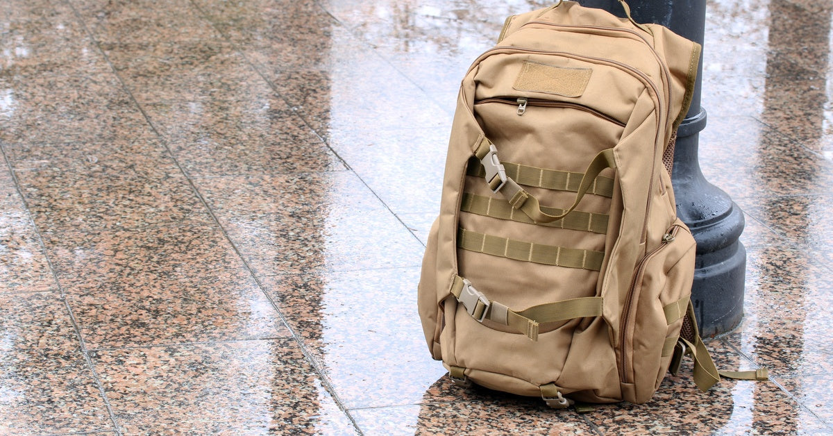 A tan tactical backpack is leaning against a black street pole on rain-soaked pavement on an urban sidewalk surface.