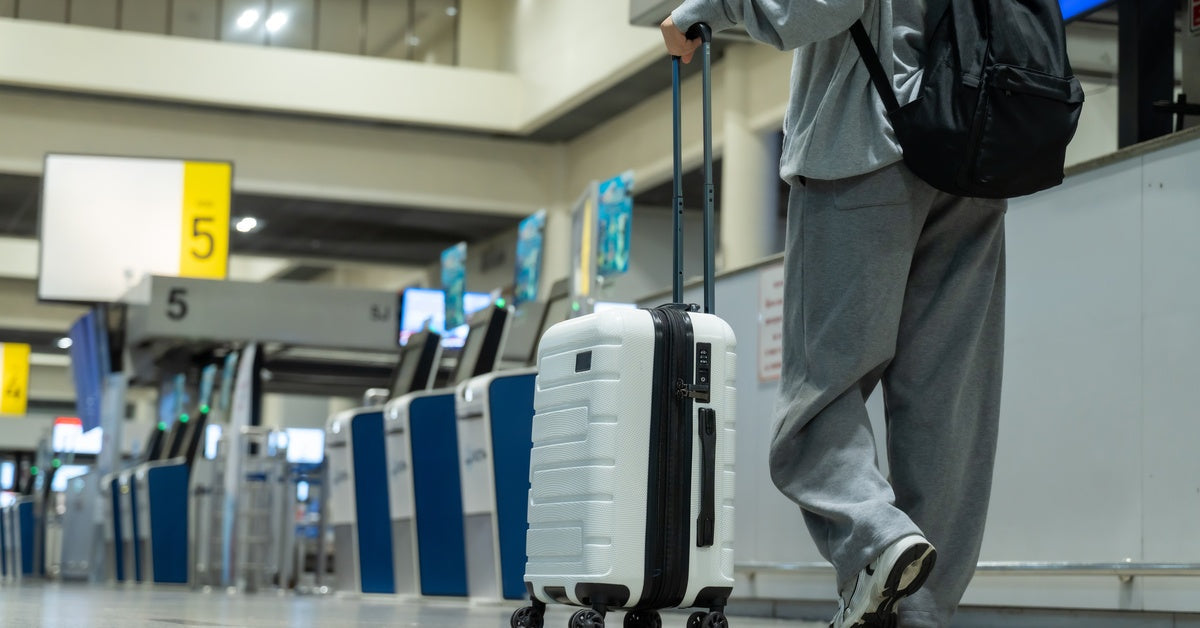 A woman walking through an airport pulls a white rolling suitcase near self-service check-in kiosks.